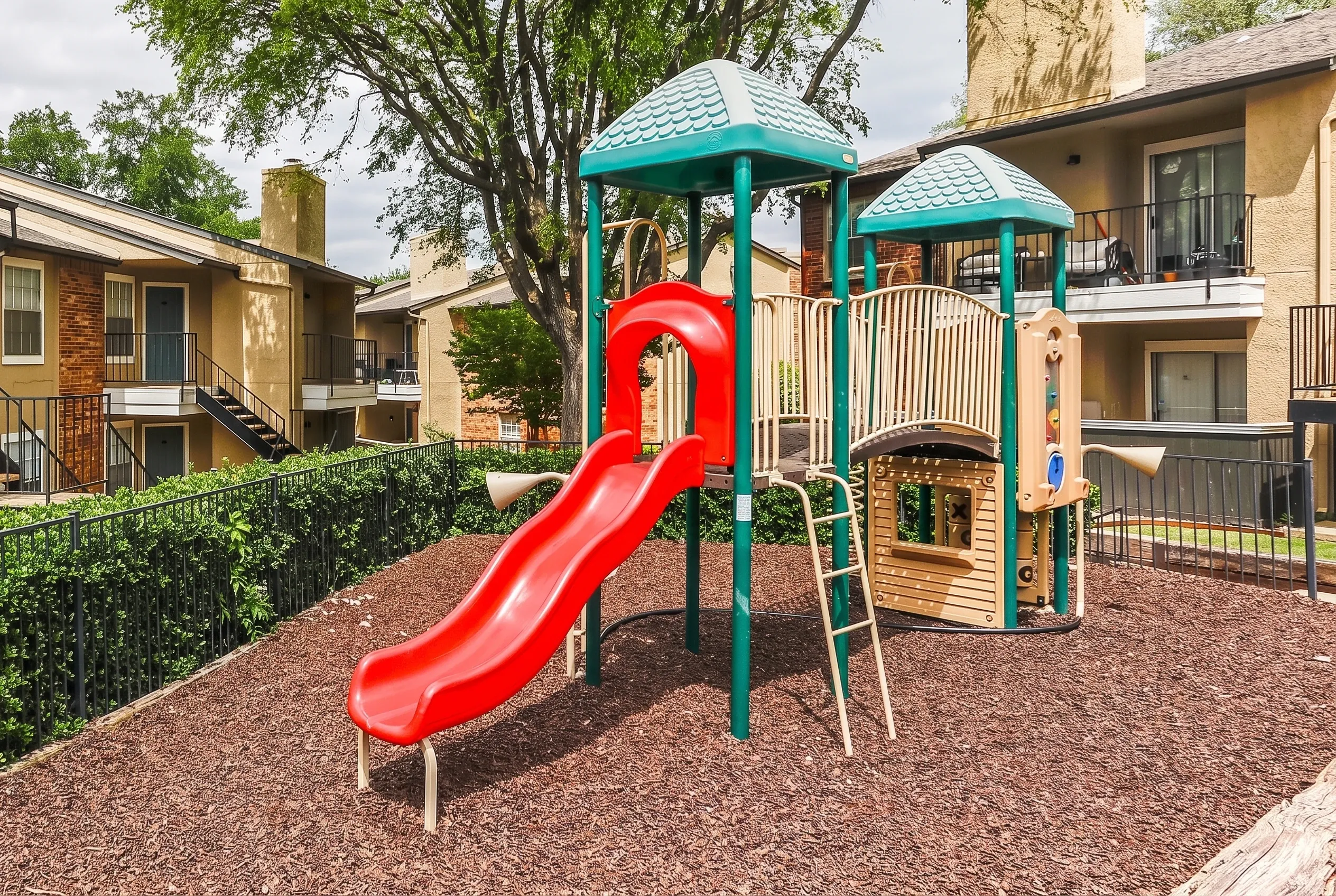 Playground with red slide, turquoise tower structures, wood-chip surfacing, residential buildings
