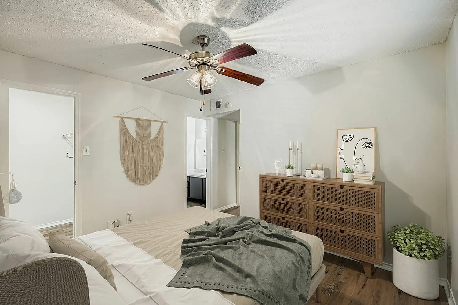 Primary bedroom, wood-style plank flooring, ceiling fan with light, wood dresser, macramé wall hanging