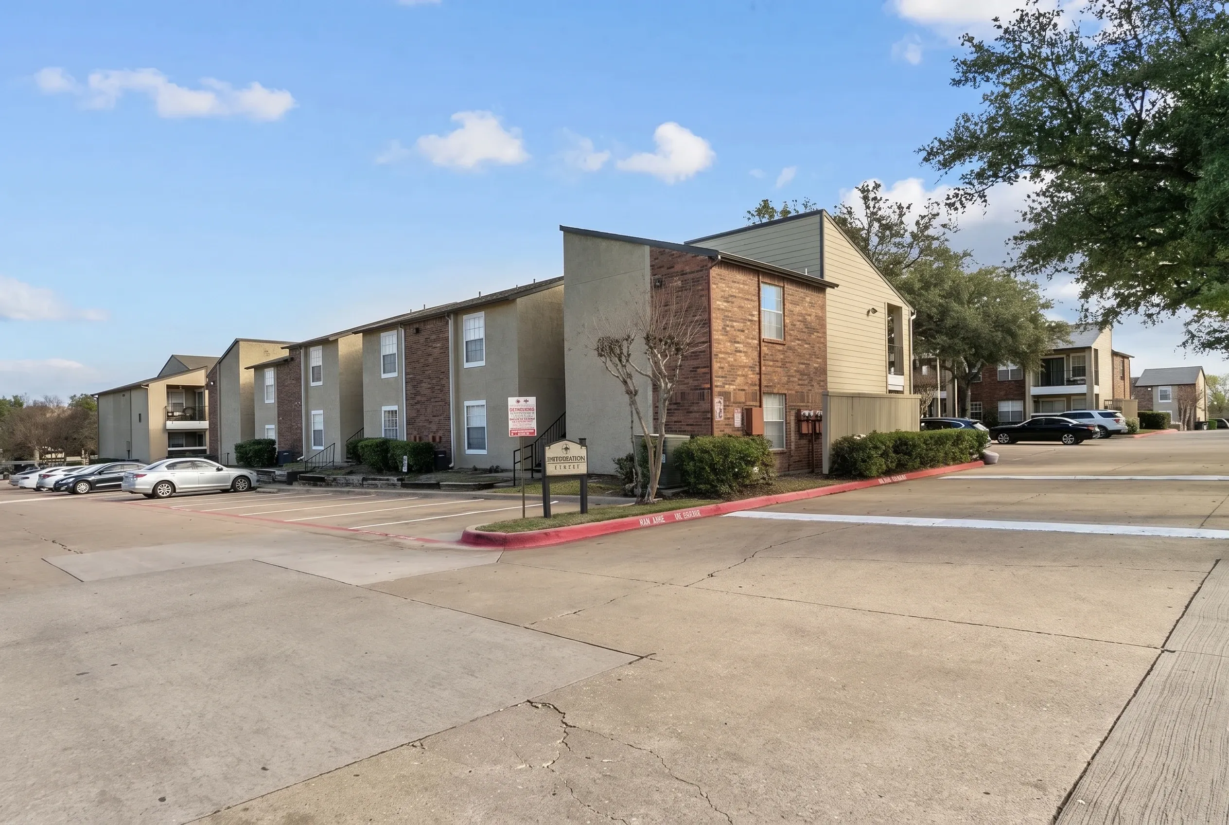 Multi-unit apartment complex with brick and tan siding, covered entry areas, paved parking lot, landscaping