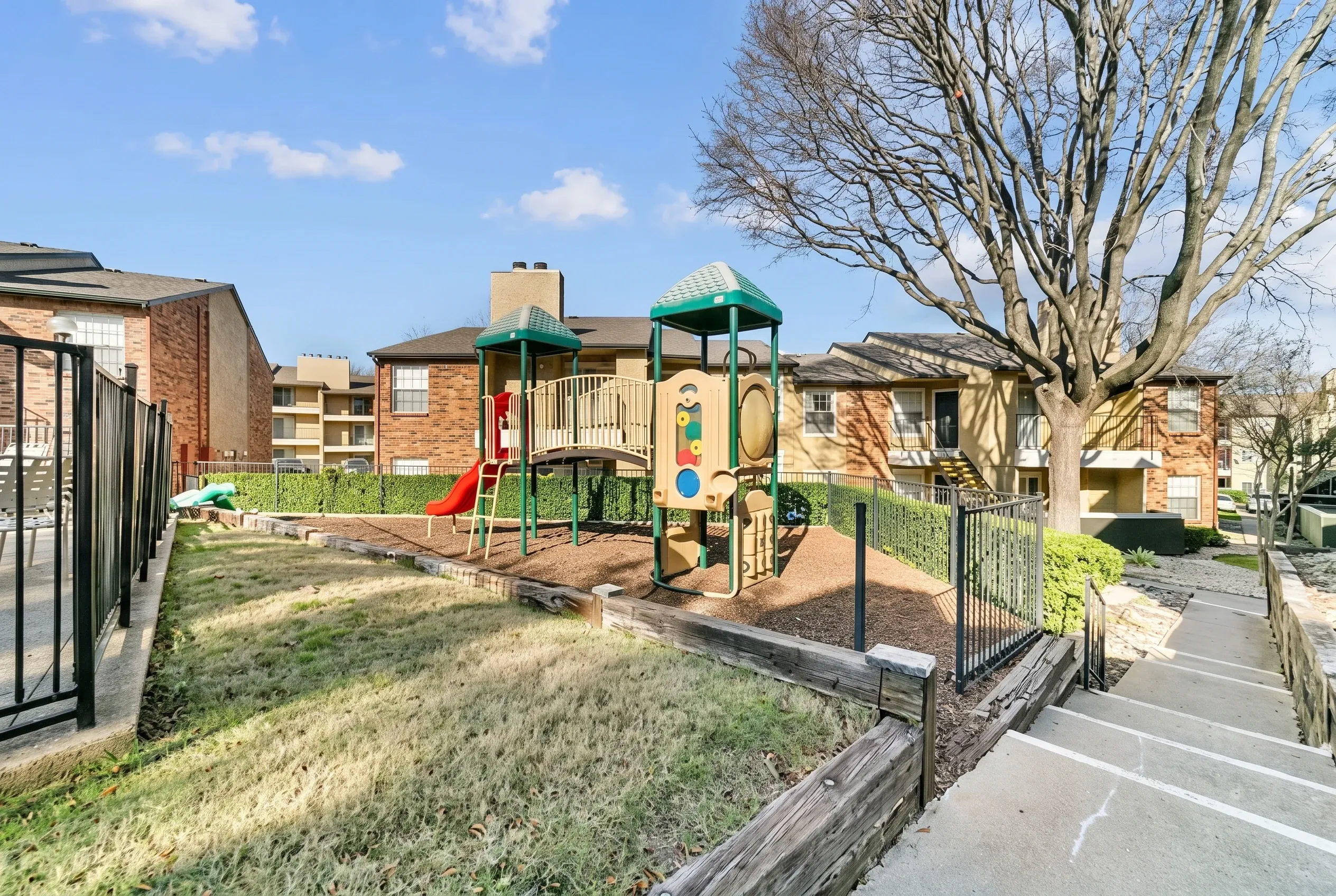 Playground area, green tower slide structure, metal fencing, brick buildings, bare tree