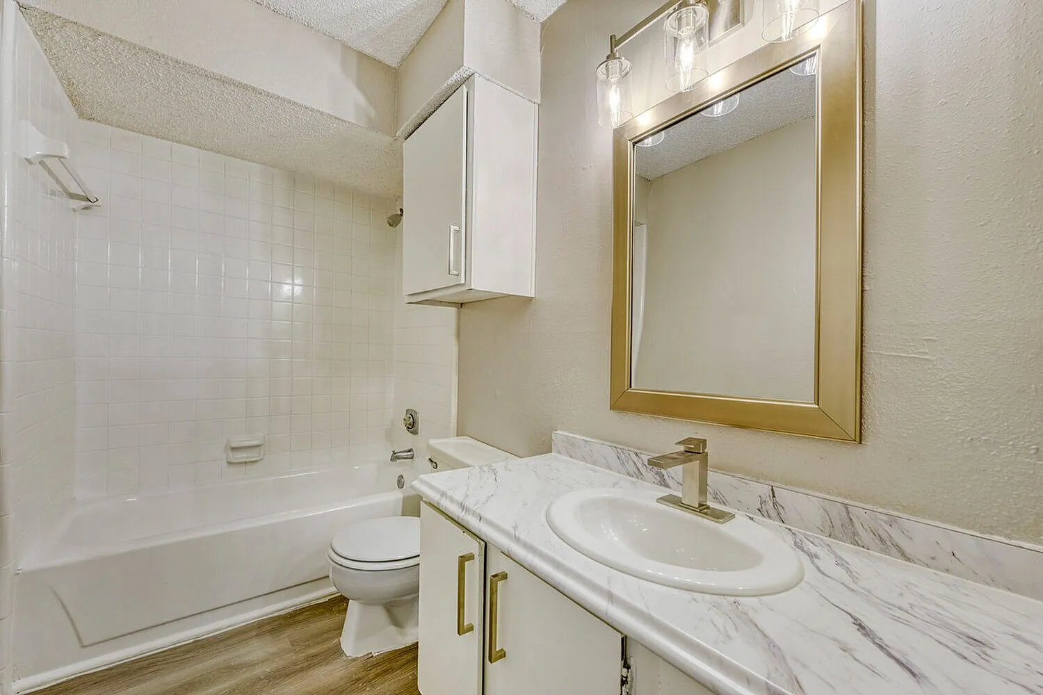 Bathroom, white vanity with marble countertop, oval sink, brass fixtures, large framed mirror, bathtub with shower, wood-style plank flooring