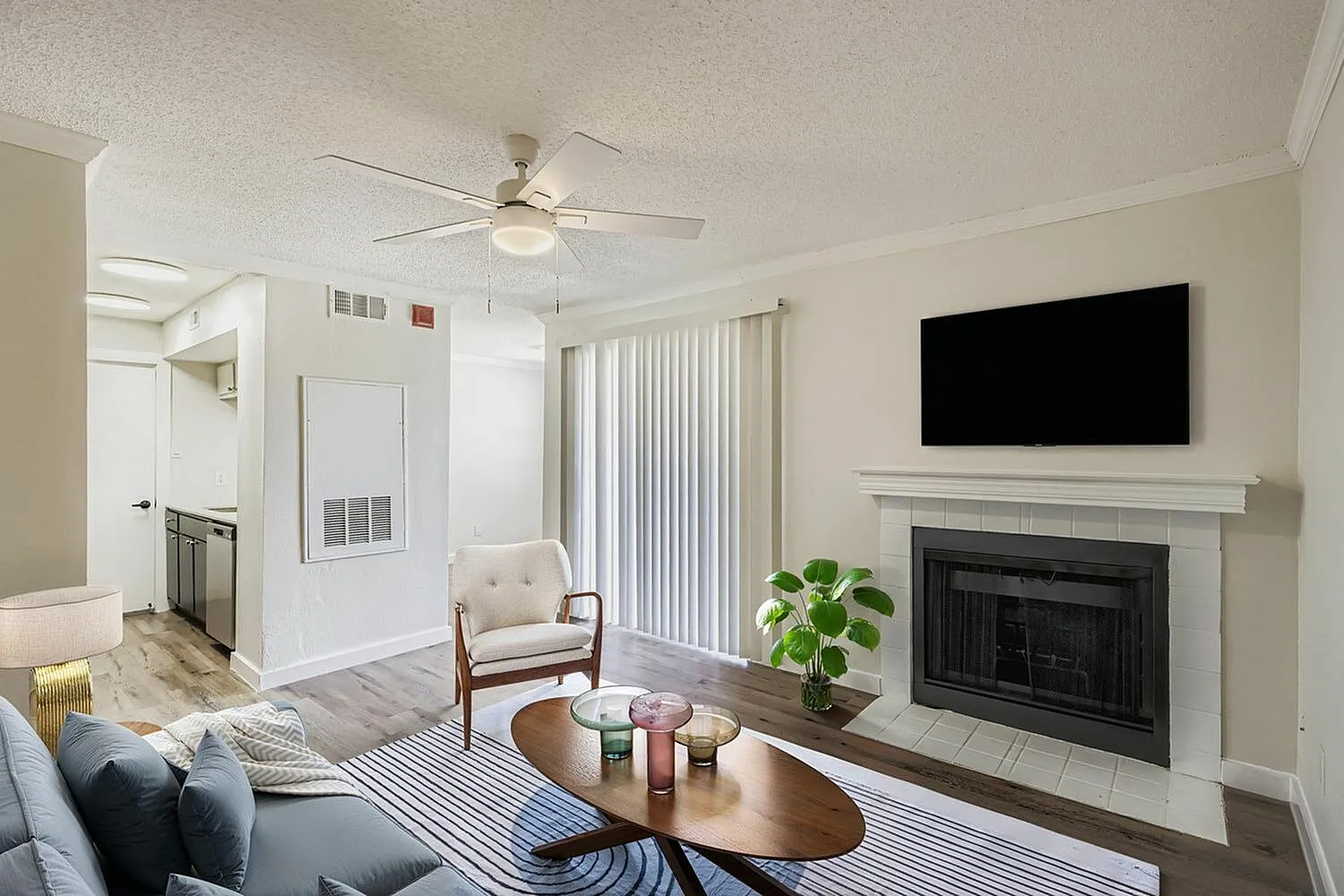 Living room, wood-style plank flooring, ceiling fan, fireplace with TV above, vertical blinds