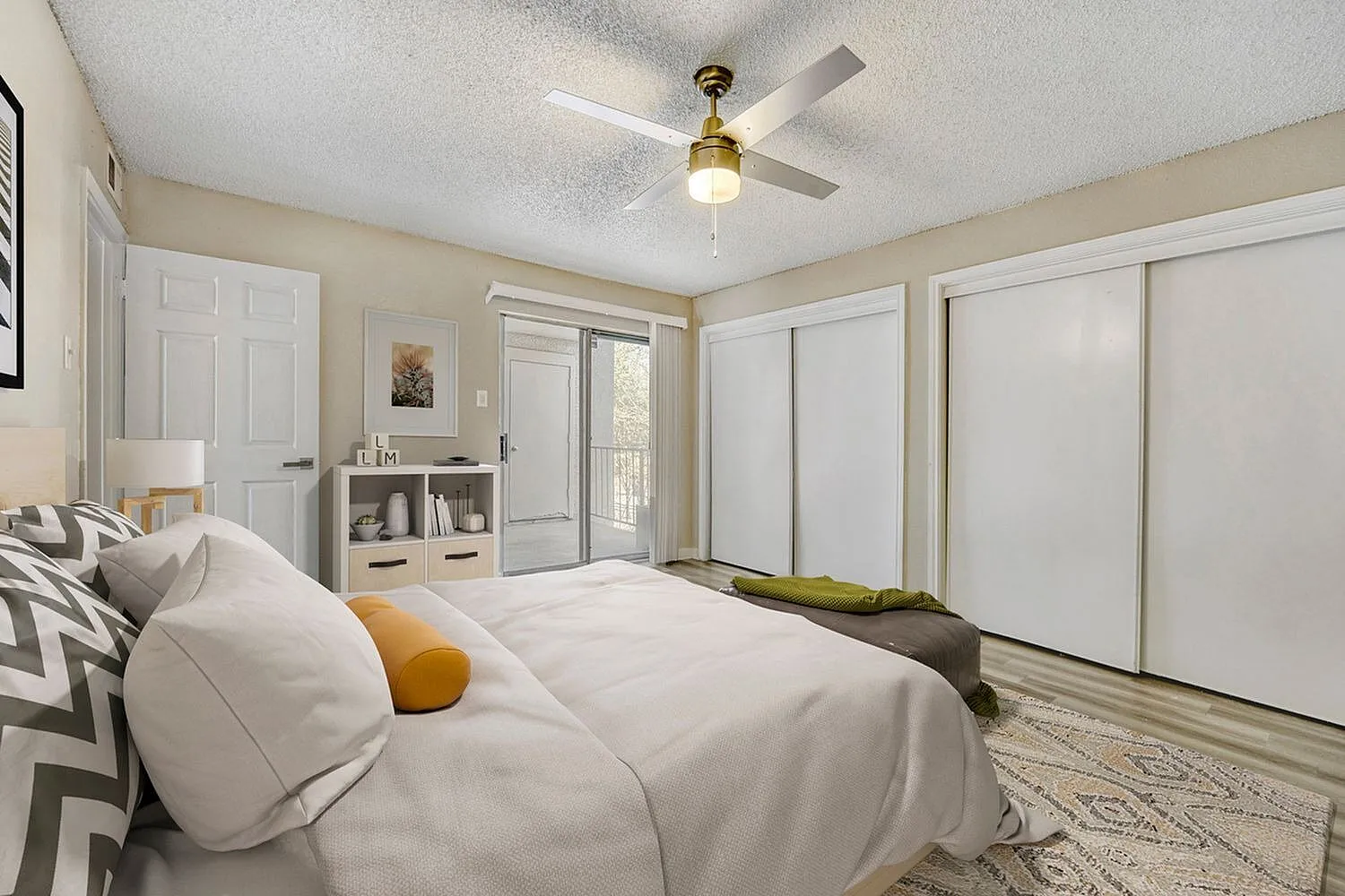 Primary bedroom, white bedding with accent pillows, ceiling fan, wood-style plank flooring, glass doors, sliding closet doors