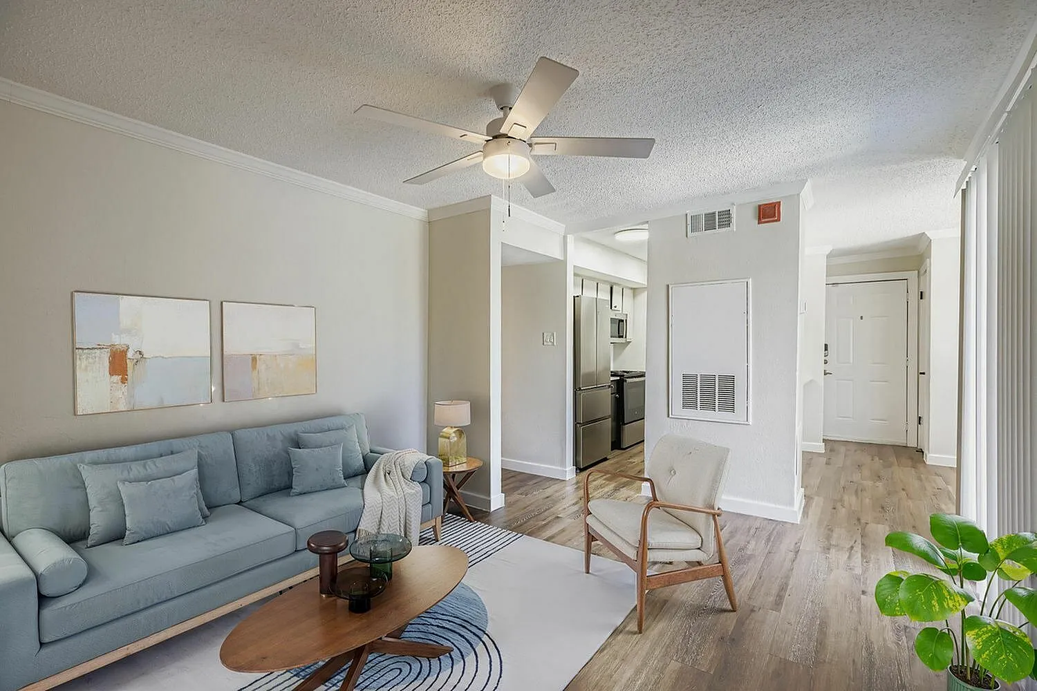 Living room with light blue sofa, wood-style plank flooring, ceiling fan, dining area and kitchen visible