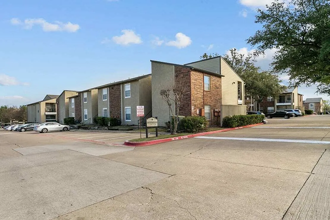 Multi-unit apartment complex with brick and tan siding, covered entry areas, paved parking lot, landscaping