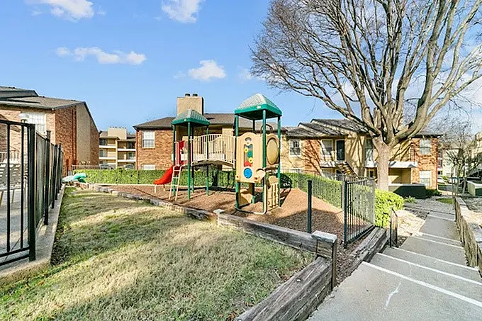 Playground area, green tower slide structure, metal fencing, brick buildings, bare tree