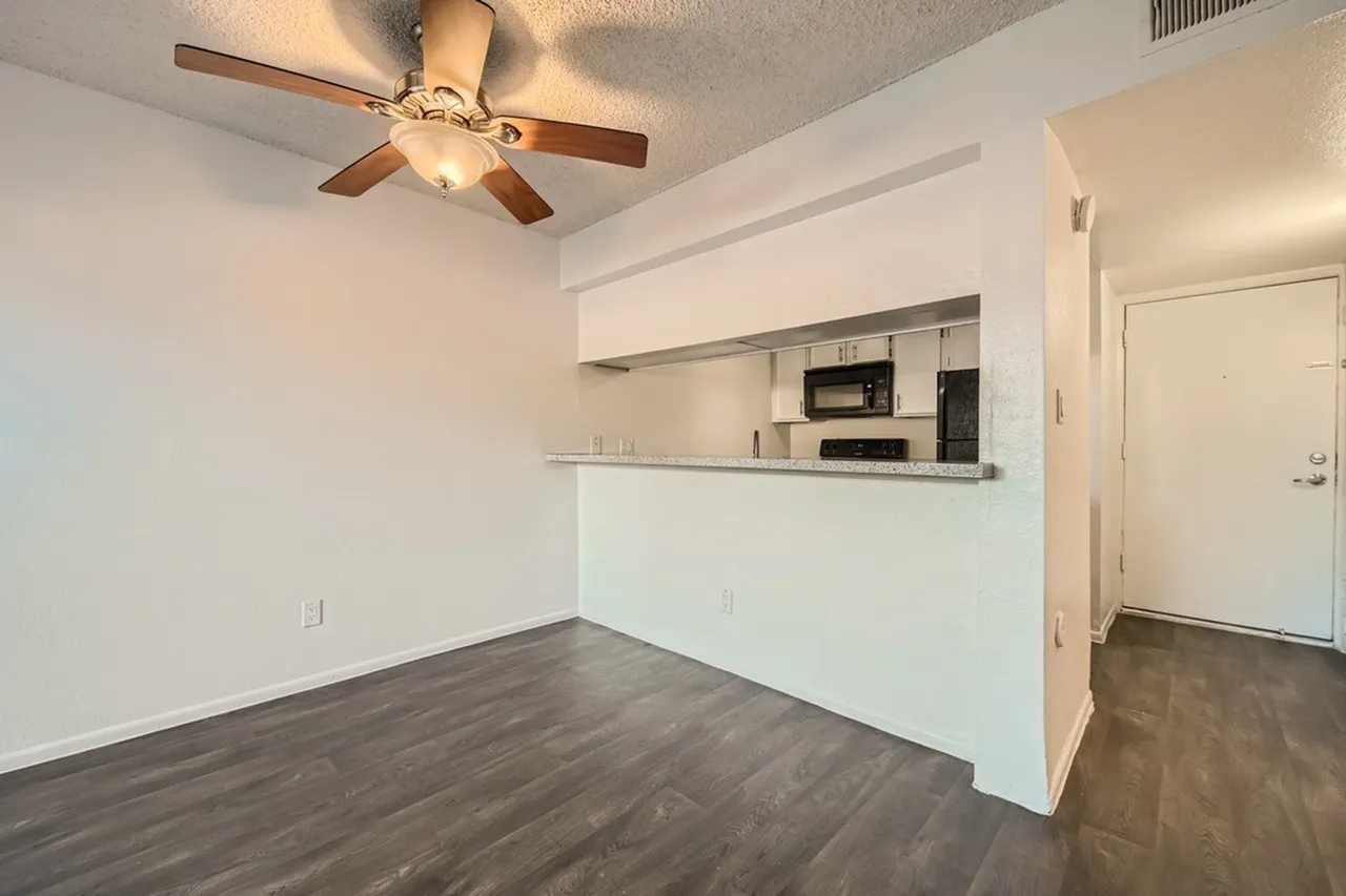 A bright, open concept living and kitchen space featuring a white kitchen counter with stainless steel appliances, dark wood-look flooring, white walls, a ceiling fan with wood blades, and a doorway leading to additional rooms