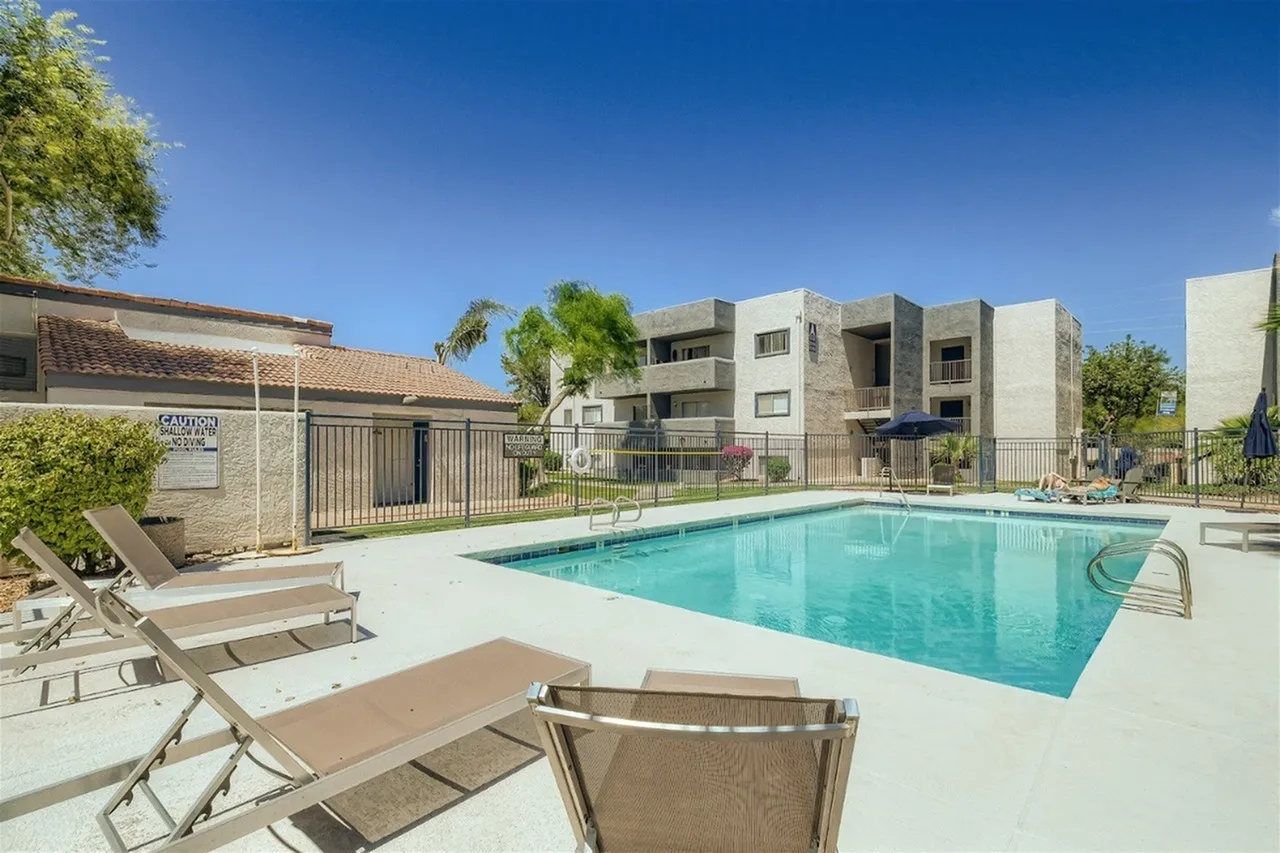 A sparkling turquoise swimming pool with lounge chairs in the foreground, surrounded by a concrete deck. A gated entry area and multi-story residential buildings with tan and stone finishes are visible in the background under a clear blue sky with green landscaping throughout the community.