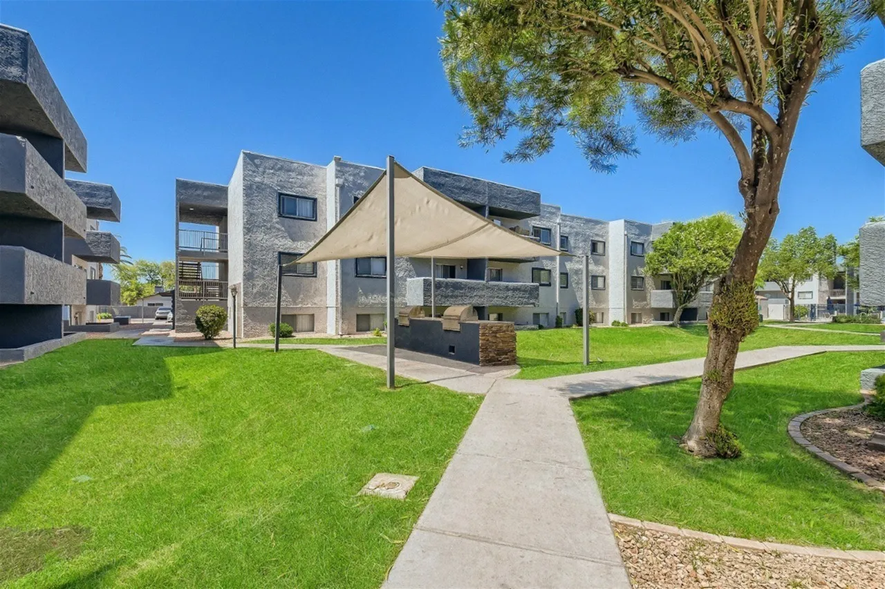 Modern apartment complex courtyard with green lawn, concrete walkway, shade sails, mature tree, and contemporary stone-and-tan residential buildings in Mesa