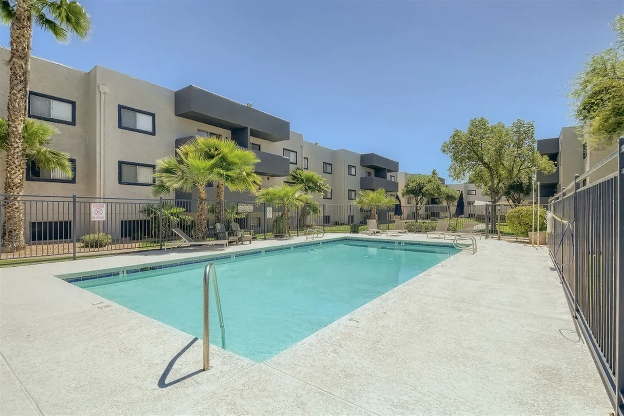 Resort-style swimming pool with turquoise water surrounded by concrete decking and metal railings, with modern multi-story residential buildings featuring beige and gray facades, palm trees, and landscaping in the background under a clear blue sky