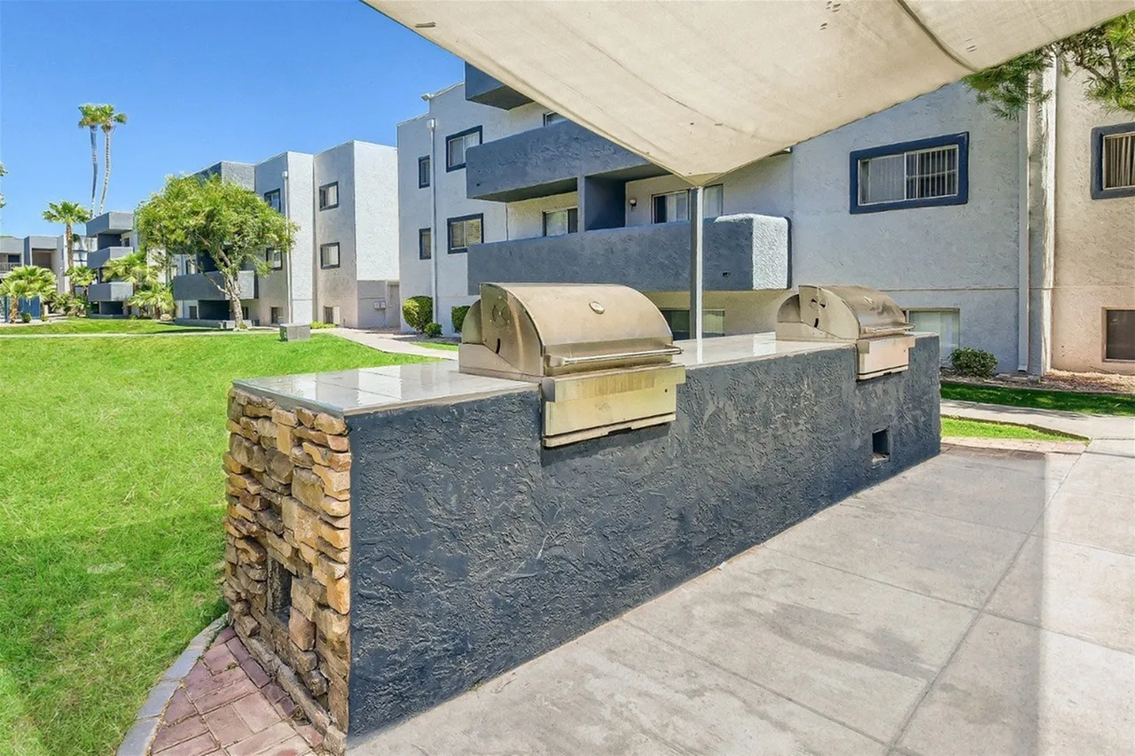 A modern outdoor kitchen area featuring a built-in barbecue grill and stainless steel cooking appliances set into a dark stone countertop with natural stone accent wall, shaded by a large beige awning, with green lawn and contemporary residential buildings visible in the background under blue sky