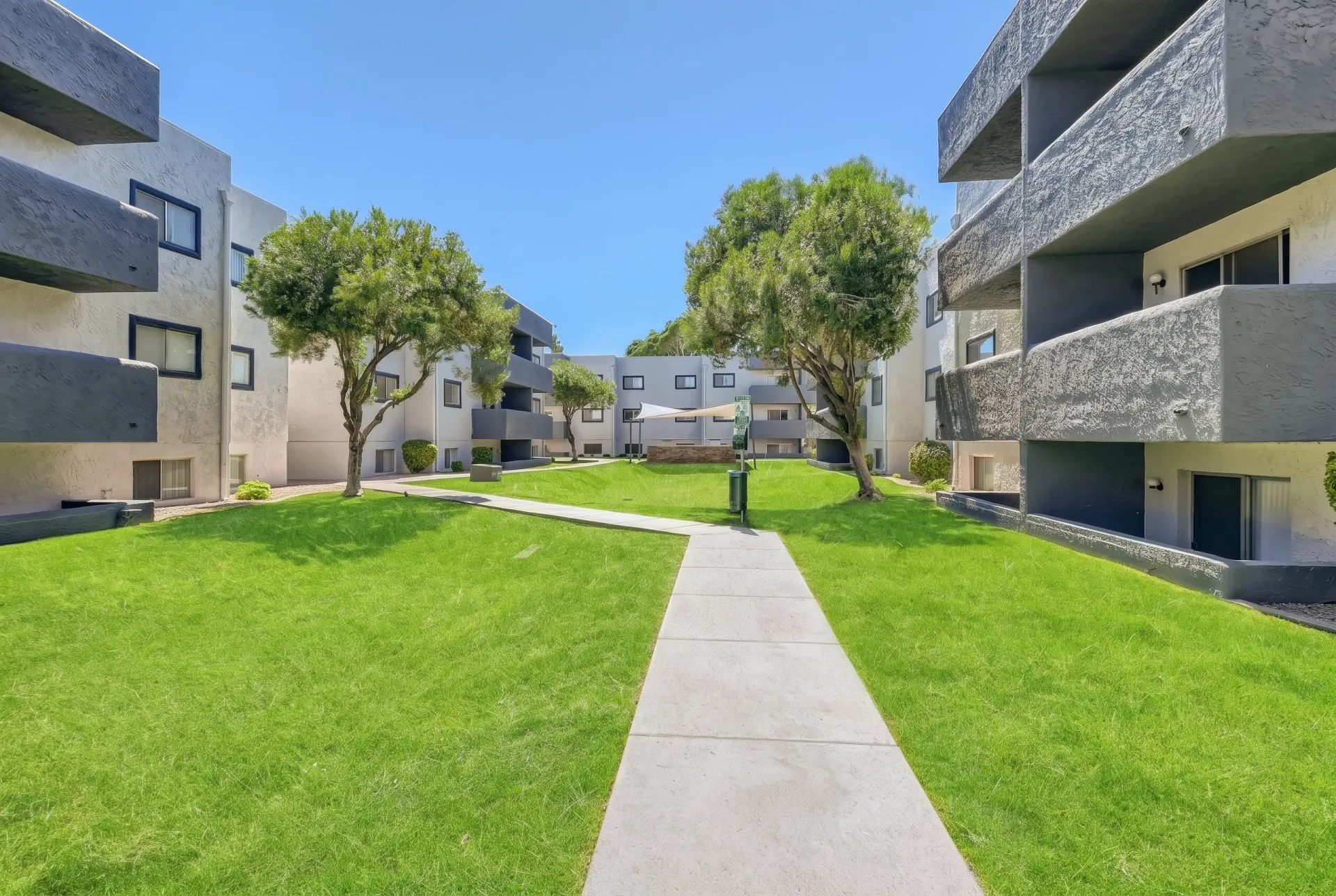 Modern residential complex with multi-story apartment buildings featuring concrete and yellow exterior panels, surrounding a spacious green lawn with two mature trees and a concrete pathway down the center under clear blue sky
