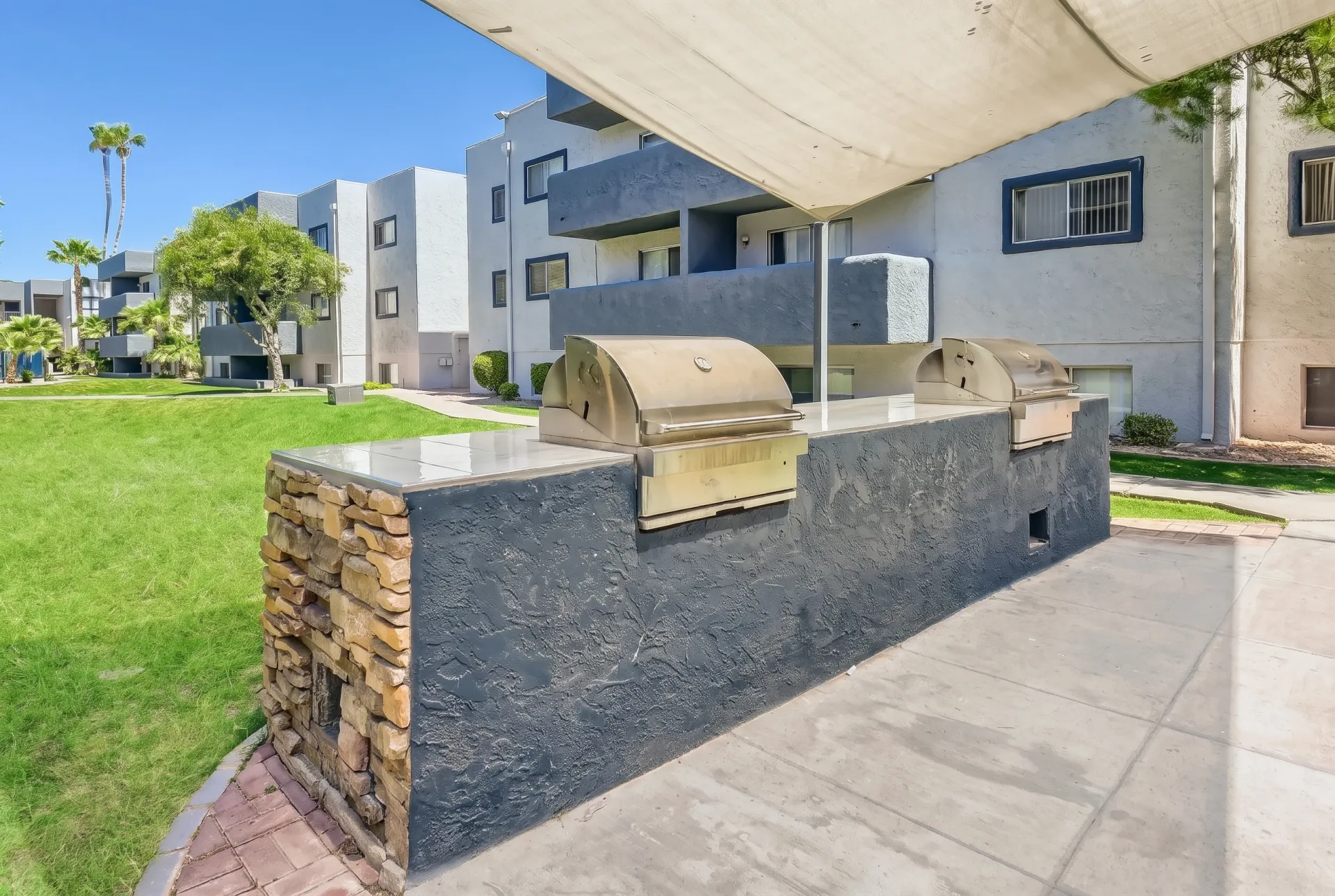 A modern outdoor kitchen area featuring a built-in barbecue grill and stainless steel cooking appliances set into a dark stone countertop with natural stone accent wall, shaded by a large beige awning, with green lawn and contemporary residential buildings visible in the background under blue sky