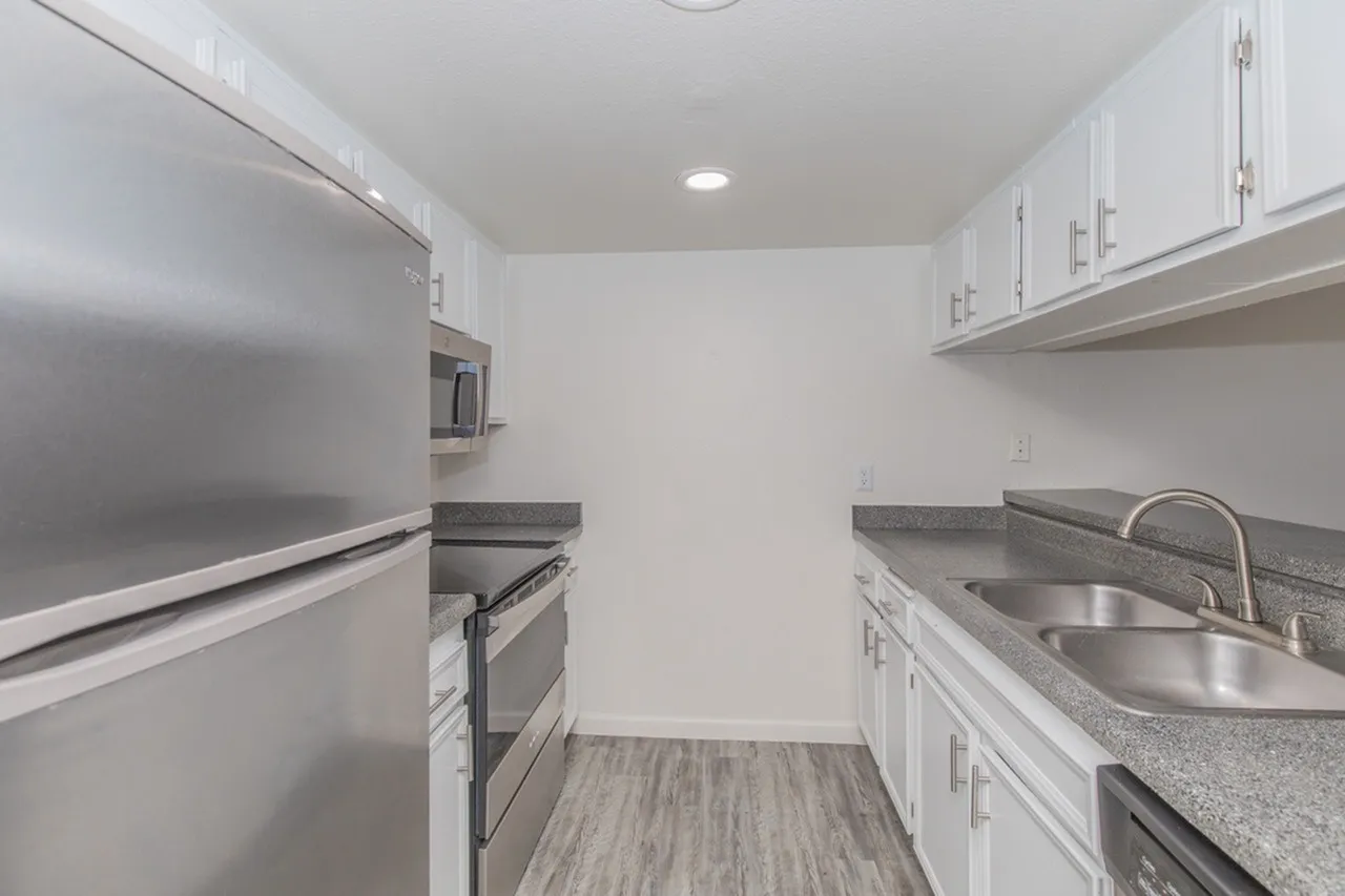 A narrow galley-style kitchen featuring white cabinetry on both sides, stainless steel appliances including a refrigerator and range, gray stone countertops with an undermount double sink, and wood-look flooring
