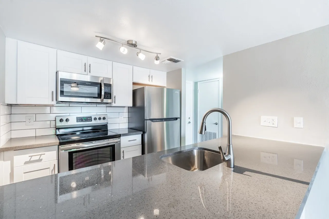 A modern kitchen featuring white cabinetry, a large center island with gray countertop, stainless steel appliances including a gas range and microwave, a refrigerator with ice maker, and a pull-down faucet over a sink. Track lighting illuminates the space and a doorway is visible to the right.