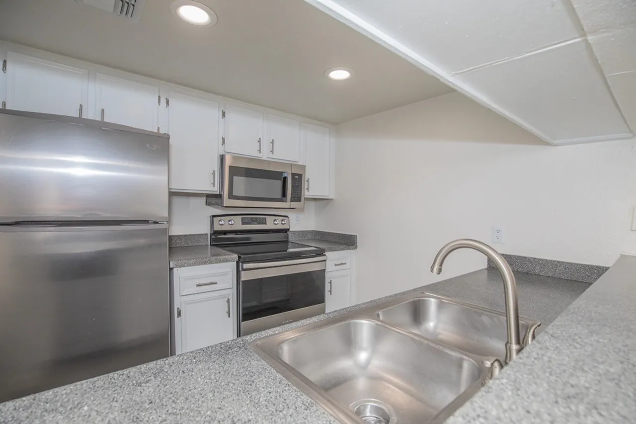 A contemporary kitchen featuring white cabinetry, stainless steel refrigerator, range, and microwave, gray quartz countertops, a double stainless steel sink with a curved faucet, recessed ceiling lights, and a window above the sink