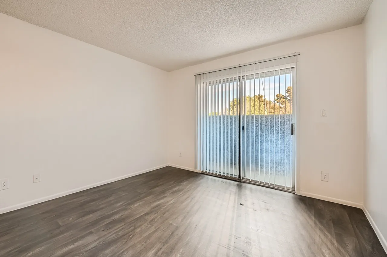 An empty apartment interior featuring light beige walls, a cream-colored ceiling, dark wood-look laminate flooring, and a large sliding glass door with vertical blinds leading to a balcony or patio area with an outdoor view