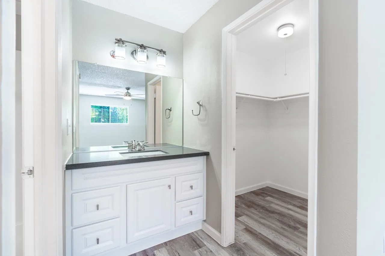 A bright, modern bathroom featuring a white vanity cabinet with a dark countertop, large mirror with decorative lighting fixture above, and an adjacent walk-in closet with open doorway and wood-grain flooring