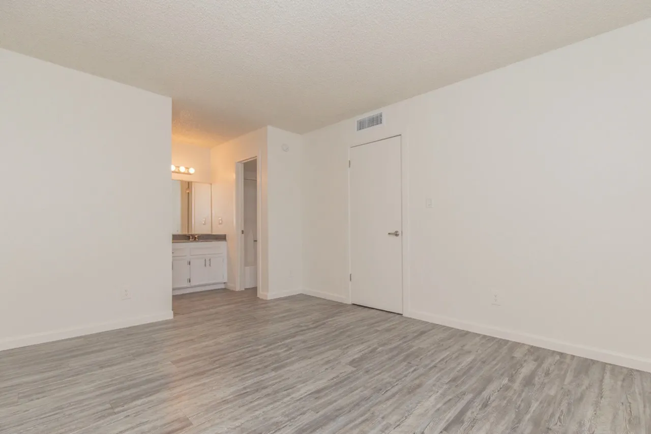 Empty bedroom with light wood plank flooring, white walls, ceiling with vent, and open doorway leading to a bathroom with vanity and mirror visible in the background