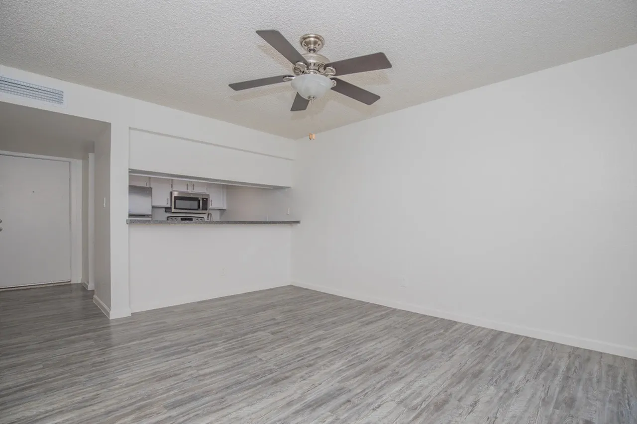 Empty apartment interior featuring a ceiling fan, white walls, gray wood-look vinyl flooring, and a kitchenette with stainless steel appliances and upper shelving along the far wall. A doorway is visible on the left side.