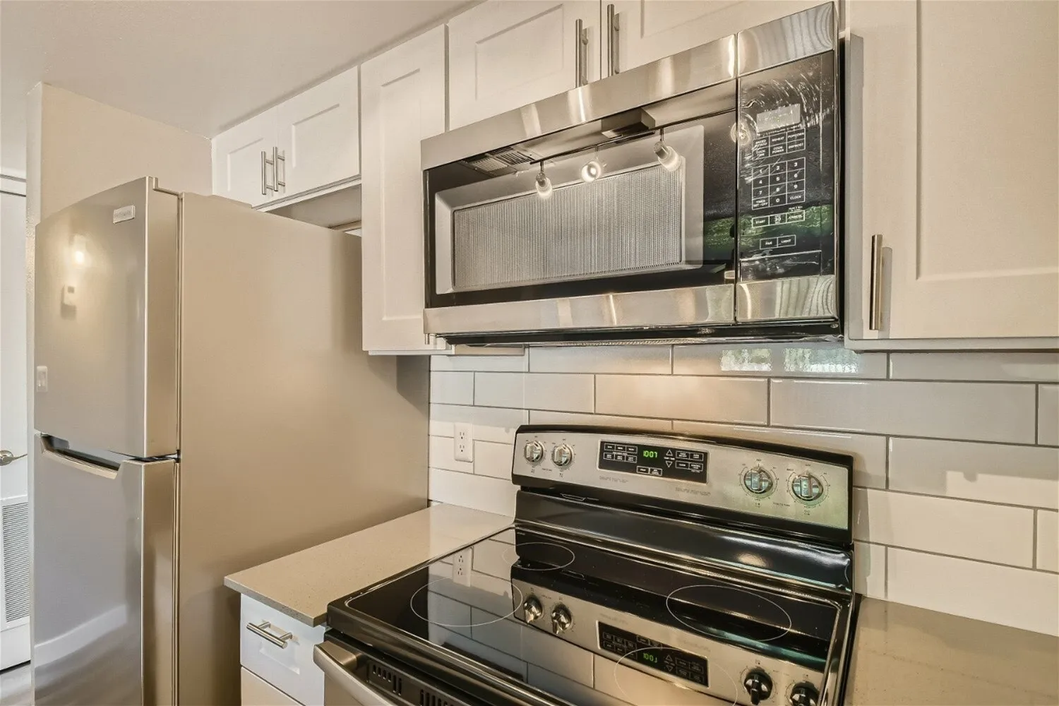A modern kitchen featuring white cabinetry, stainless steel appliances including a side-by-side refrigerator, double wall ovens, microwave, and subway tile backsplash with a light gray countertop