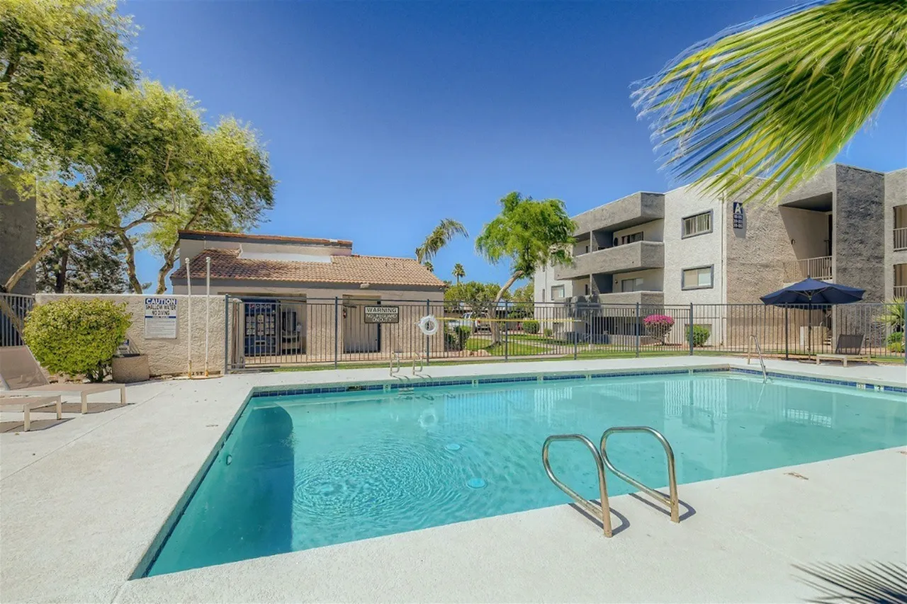 A sparkling rectangular swimming pool with turquoise water and metal ladder on the concrete deck, surrounded by a modern multi-story residential building, pool house structure, mature trees, and clear blue sky