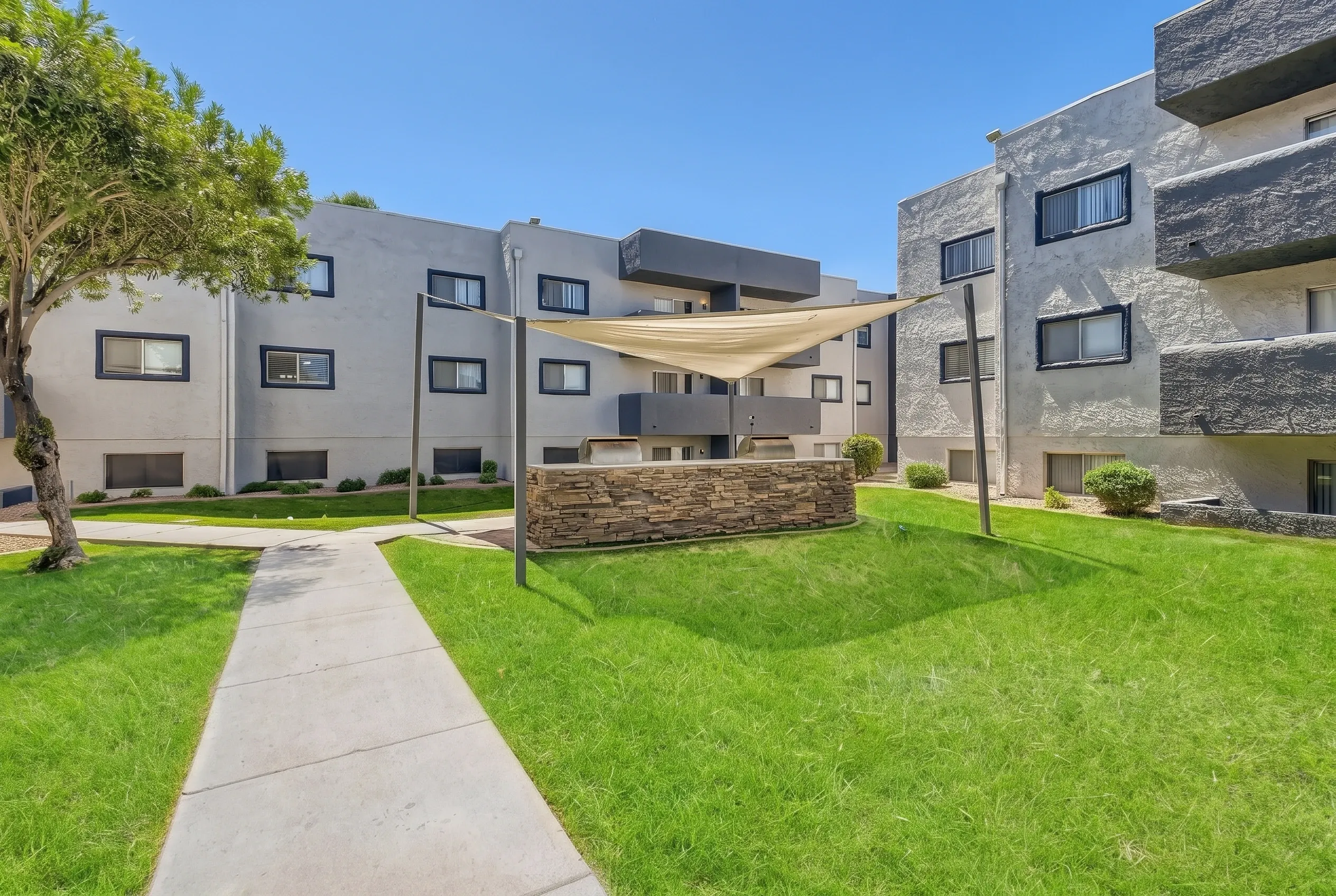 Modern multi-story residential buildings with stone and stucco exteriors surrounding a landscaped courtyard with green lawn, mature trees, concrete walkway, planter boxes, and a cream-colored shade canopy
