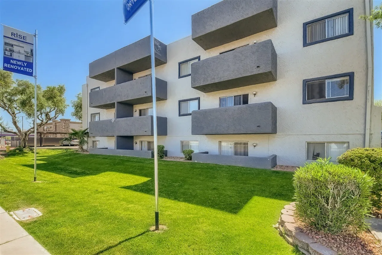 Modern multi-story residential building with stacked concrete balconies, white and grey facade, surrounded by manicured lawn, green shrubs, and a flagpole, photographed on a clear sunny day