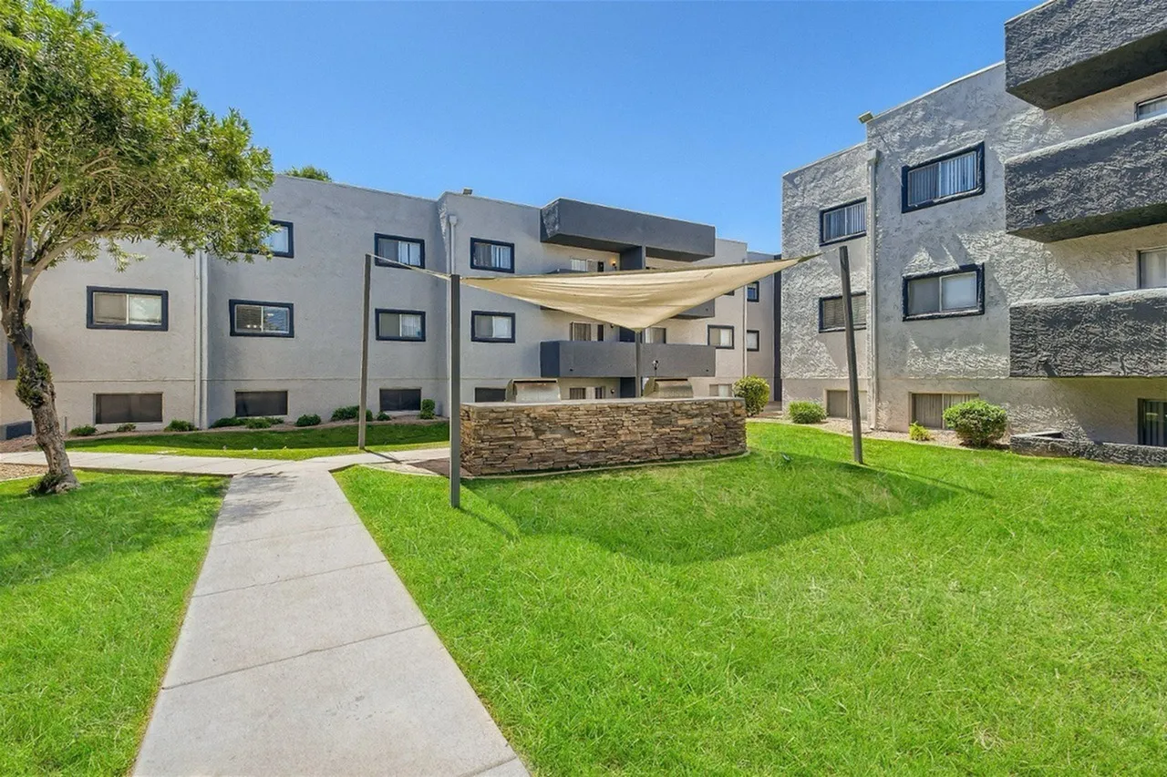 Modern multi-story residential buildings with stone and stucco exteriors surrounding a landscaped courtyard with green lawn, mature trees, concrete walkway, planter boxes, and a cream-colored shade canopy