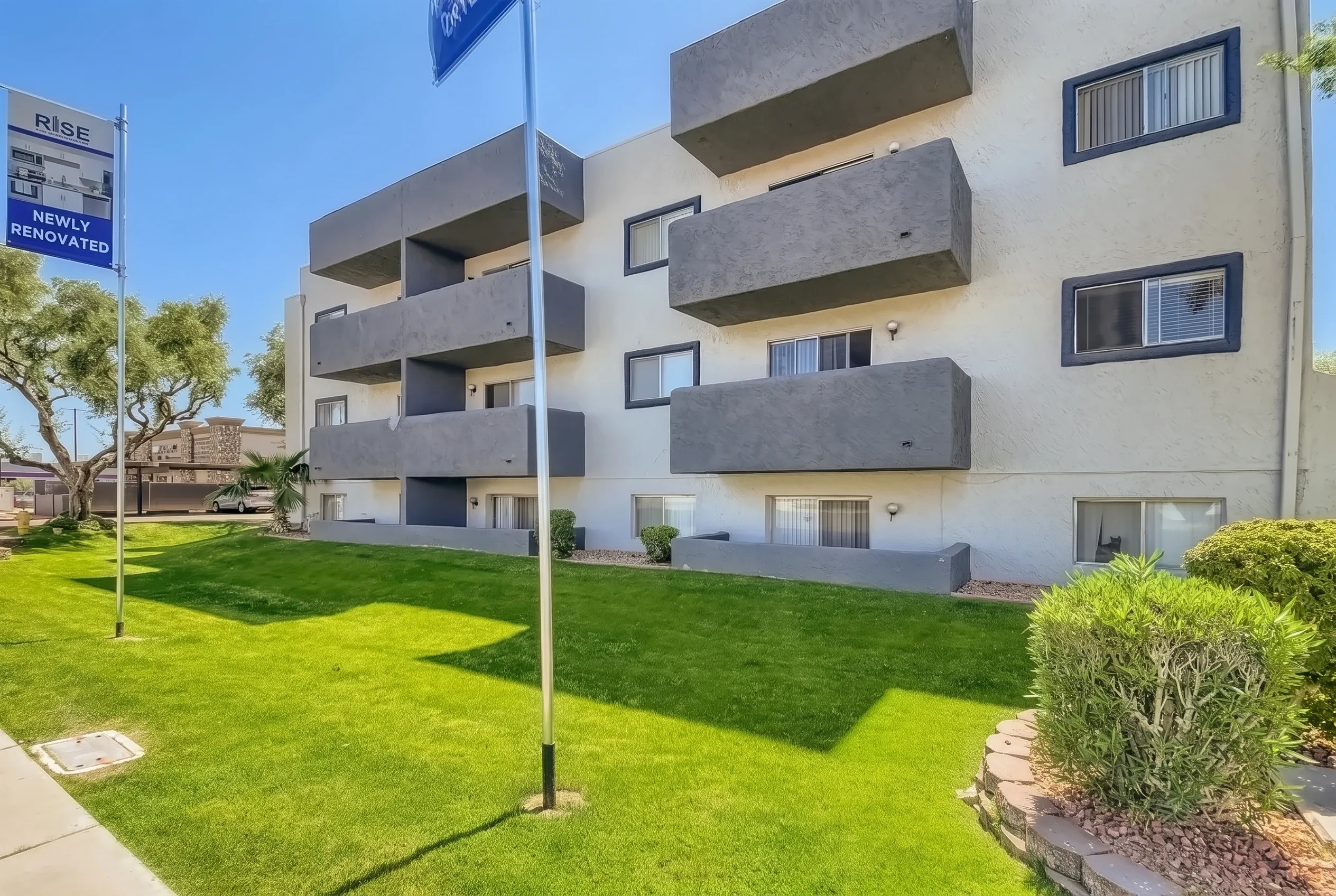 Modern multi-story residential building with stacked concrete balconies, white and grey facade, surrounded by manicured lawn, green shrubs, and a flagpole, photographed on a clear sunny day