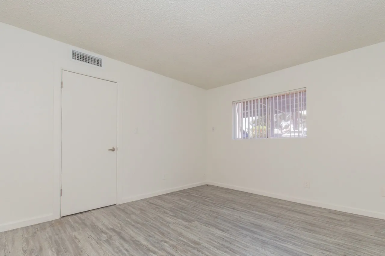 Empty bedroom with white walls, wood-look vinyl flooring, a closed white door on the left wall, a small ceiling-mounted HVAC vent, and a window with vertical blinds on the right wall letting in natural light