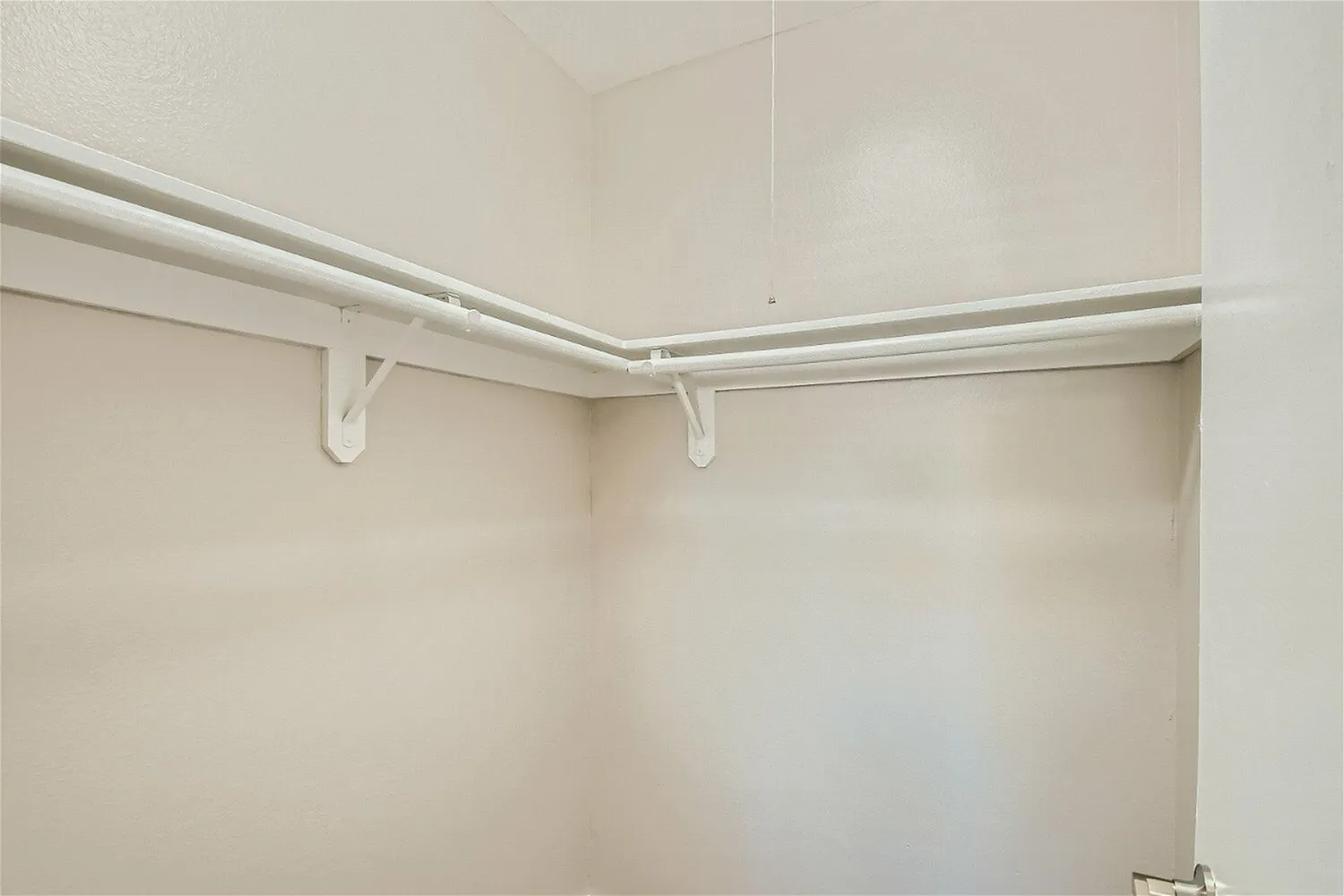 Interior view of an empty walk-in closet featuring white painted walls, a ceiling-mounted chrome hanging rod with white bracket supports, and built-in shelving space