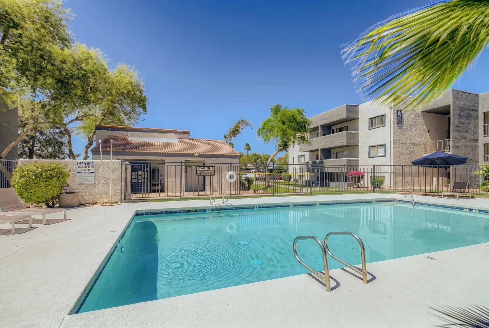 A sparkling rectangular swimming pool with turquoise water and metal ladder on the concrete deck, surrounded by a modern multi-story residential building, pool house structure, mature trees, and clear blue sky