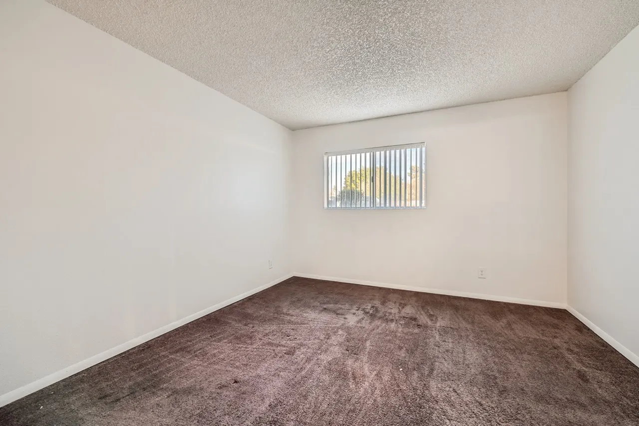 Empty bedroom with brown carpet, white walls, textured ceiling, and a window with vertical blinds on the far wall