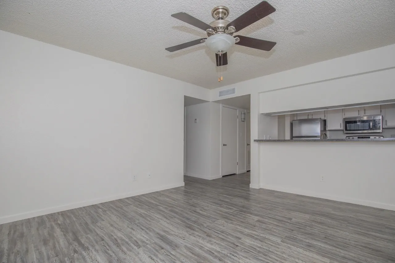 Empty apartment interior featuring gray wood-look flooring, white walls, a brushed nickel ceiling fan with light fixture, open doorway leading to a hallway, and stainless steel kitchen shelving visible on the right wall