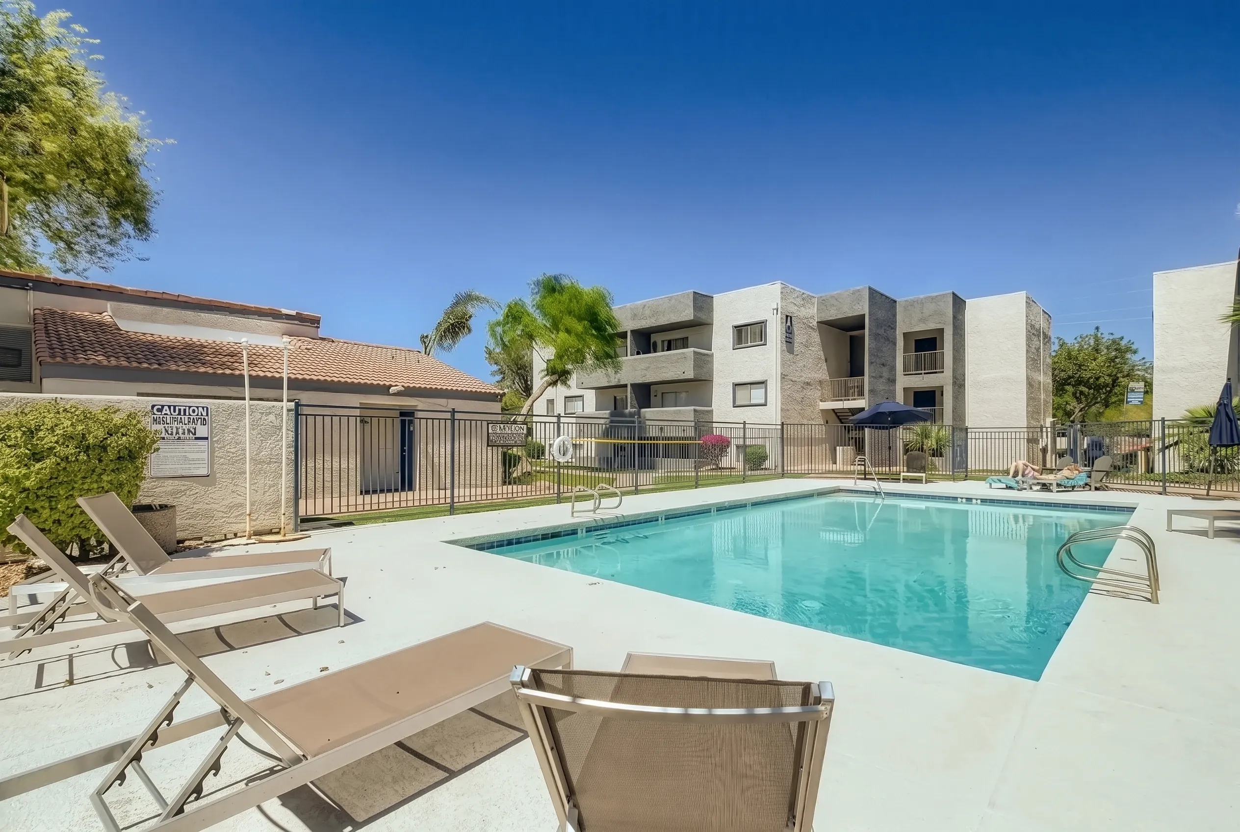 A sparkling turquoise swimming pool with lounge chairs in the foreground, surrounded by a concrete deck. A gated entry area and multi-story residential buildings with tan and stone finishes are visible in the background under a clear blue sky with green landscaping throughout the community.