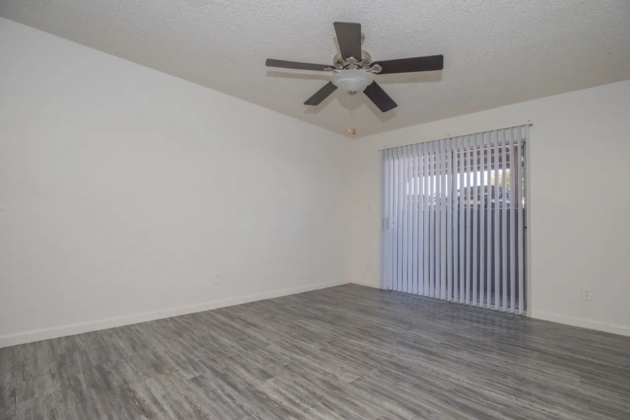 A spacious empty bedroom with light wood-look flooring, white walls, a black ceiling fan with light fixture, and vertical blinds covering a sliding glass door on the right side