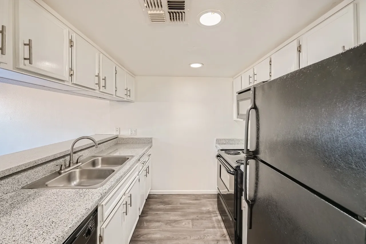 A narrow galley-style kitchen with white cabinetry lining both sides, featuring a double sink with chrome faucet on the left, granite countertops, stainless steel appliances including a range and refrigerator on the right, recessed and pendant lighting, and a wood-look plank floor.