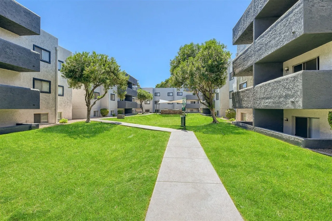 Modern residential complex with multi-story apartment buildings featuring concrete and yellow exterior panels, surrounding a spacious green lawn with two mature trees and a concrete pathway down the center under clear blue sky