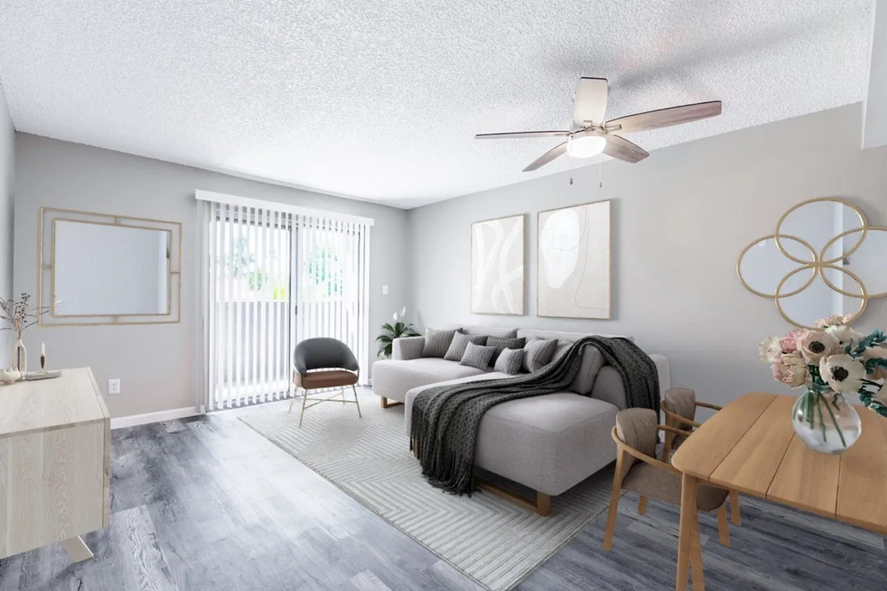 A bright, modern apartment interior featuring a gray sofa with throw pillows and a matching armchair, ceiling fan, large windows with vertical blinds, hardwood-look flooring, a round dining table with wood base, decorative wall art, fresh flowers in a vase, and neutral color palette throughout