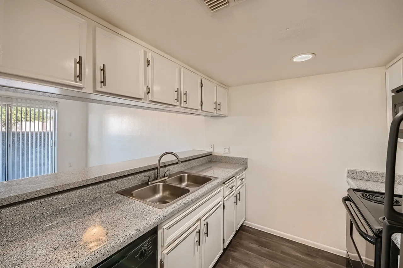 Modern kitchen featuring cream-colored cabinetry with stainless steel hardware, gray speckled granite countertops, a double stainless steel sink, and a window with vertical blinds allowing natural light. Hard wood flooring and recessed ceiling lights are visible.