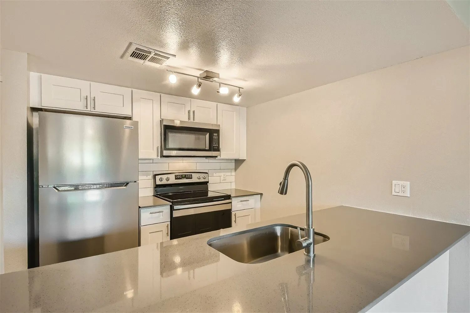 A contemporary kitchen featuring white cabinetry, stainless steel refrigerator and range, black and stainless cooktop, undermount sink with faucet, and gray quartz or granite countertops with an island in the foreground. Recessed and track lighting illuminate the space.