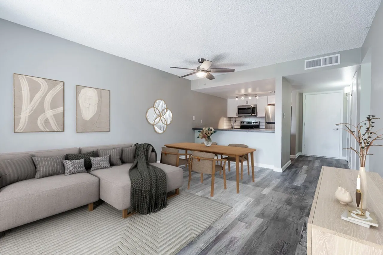 A bright, open-concept living room with a curved gray sectional sofa, wooden dining table with chairs, abstract wall art, ceiling fan, and kitchen visible in the background with stainless steel appliances