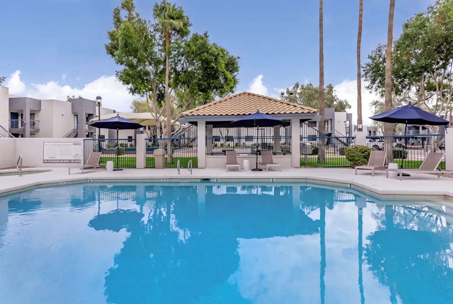 Resort-style pool with clear blue water, cabanas with blue umbrellas, lounge seating, palm trees, modern buildings