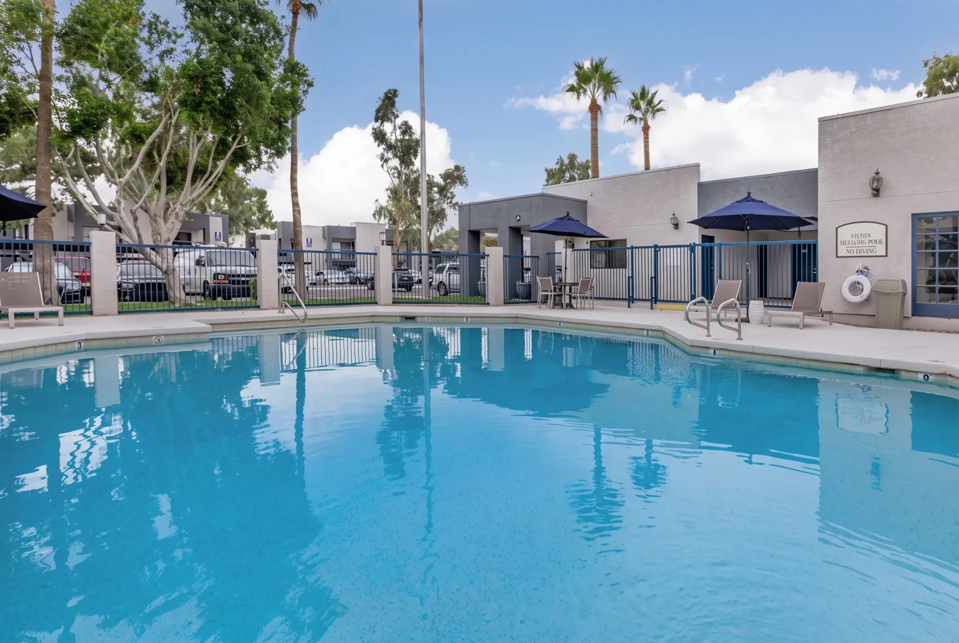 Resort-style swimming pool, blue umbrellas, lounge seating, white building structures, palm trees