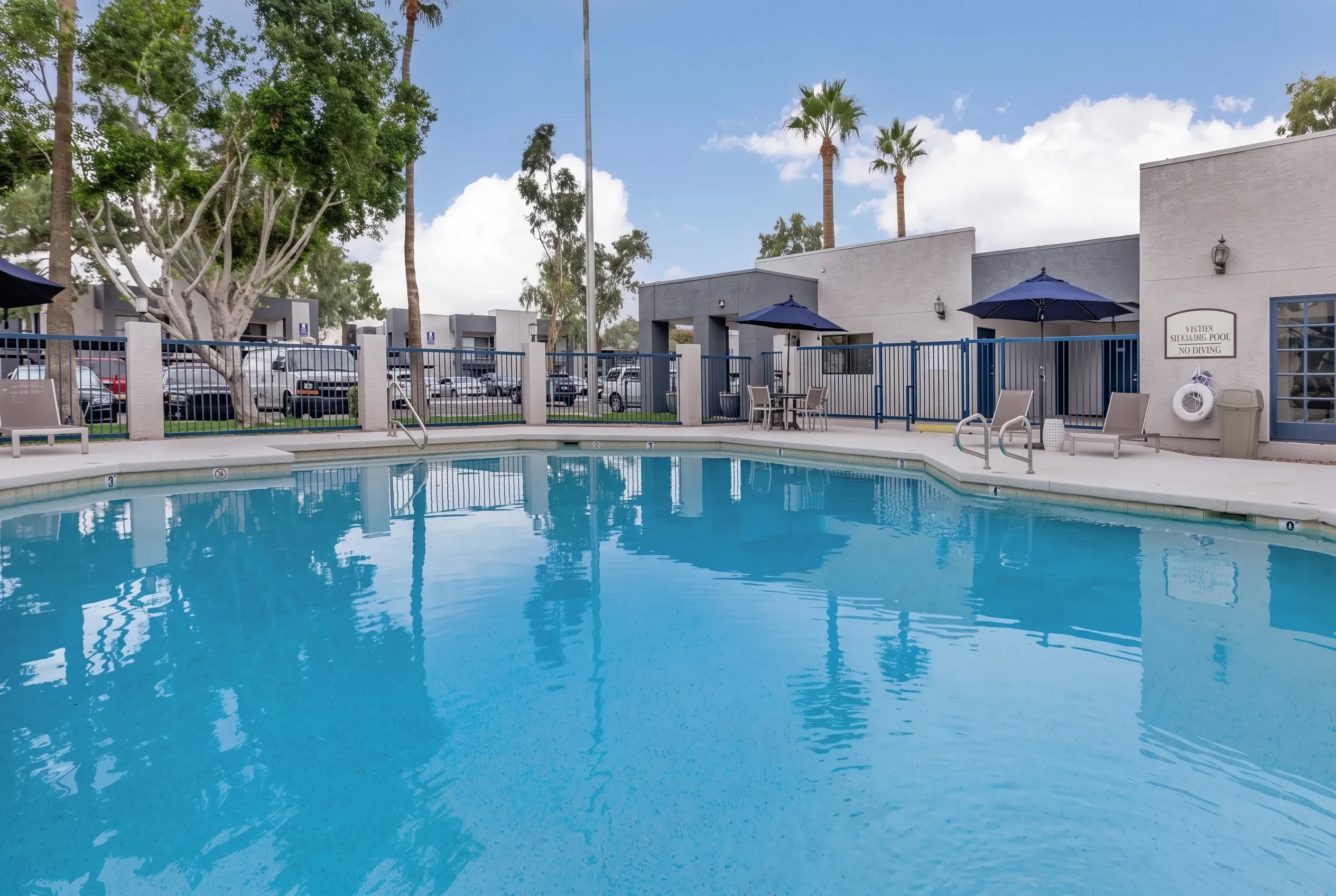 Resort-style swimming pool, blue umbrellas, lounge seating, white building structures, palm trees