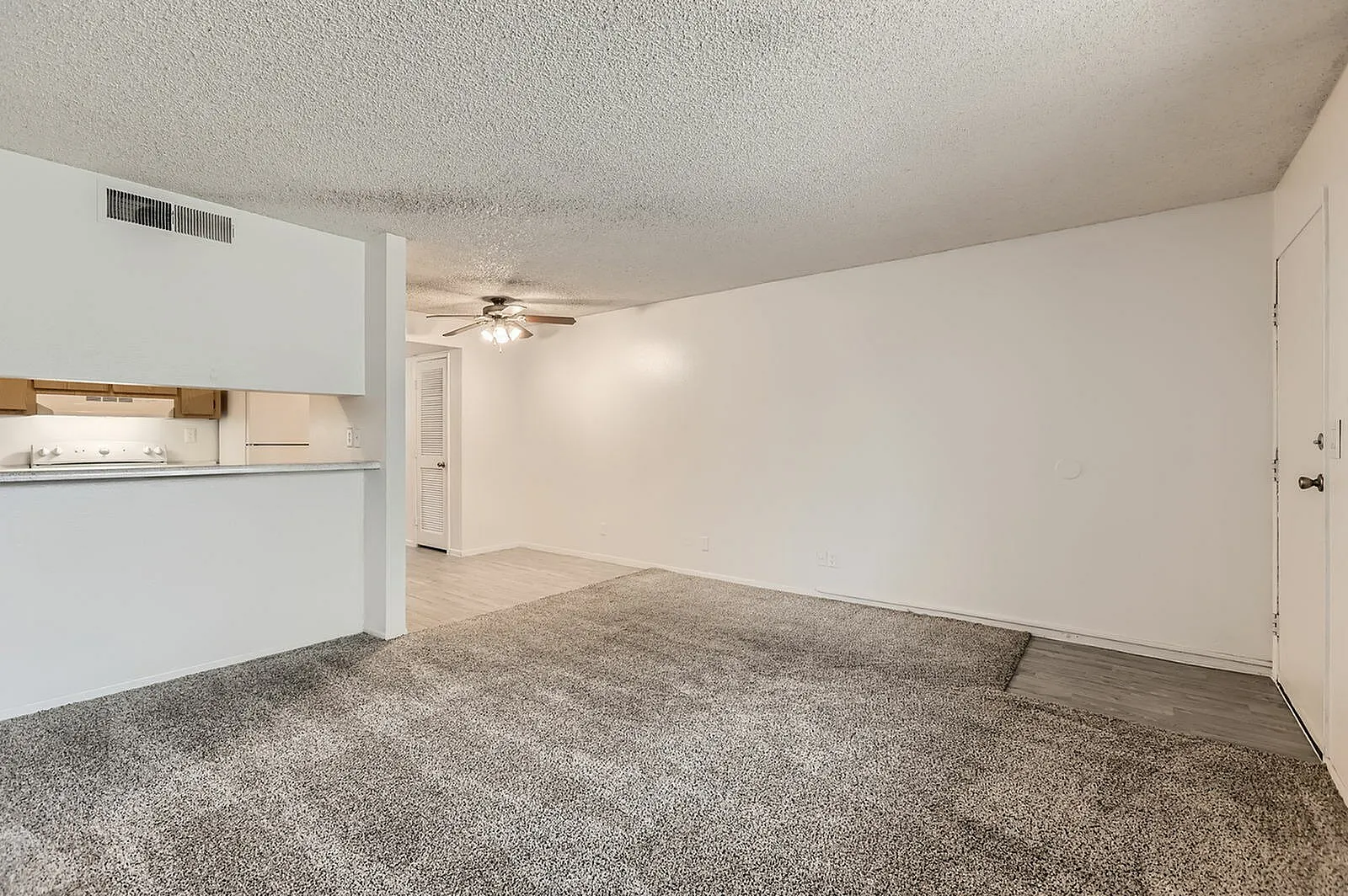 Living room with gray carpet flooring, ceiling fan, open kitchen with white cabinetry, entry door