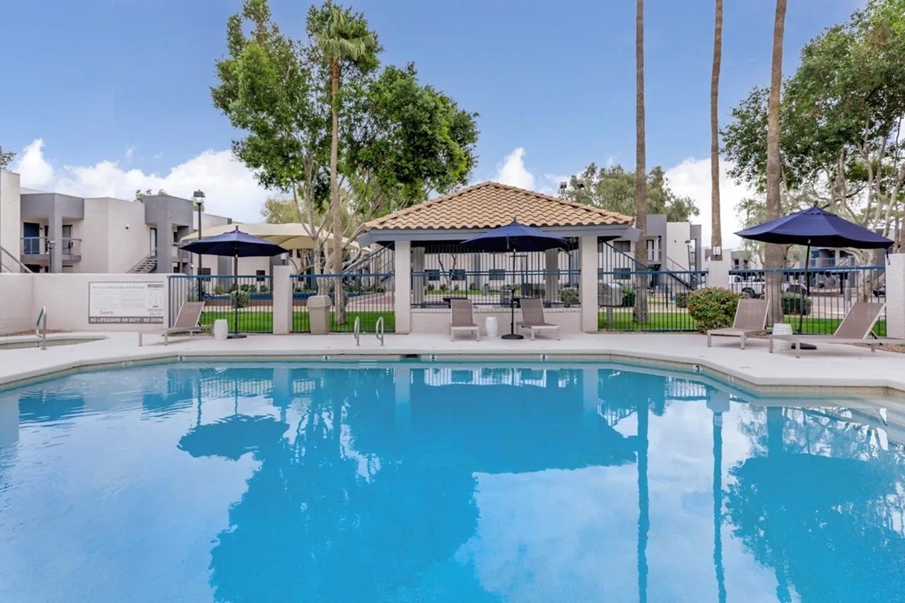 Resort-style pool with clear blue water, cabanas with blue umbrellas, lounge seating, palm trees, modern buildings