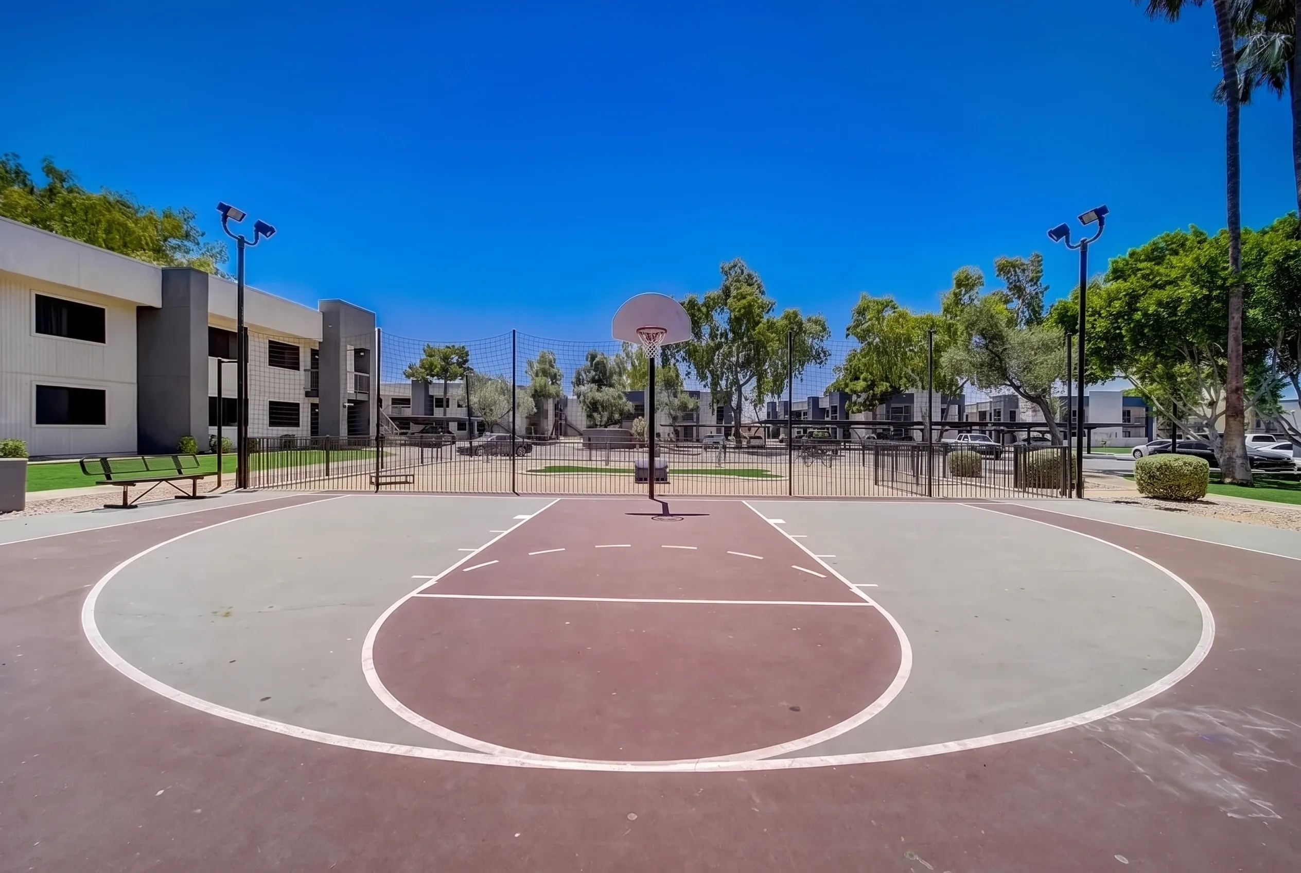 Outdoor basketball court, red and gray surface, white line markings, apartment building, palm trees