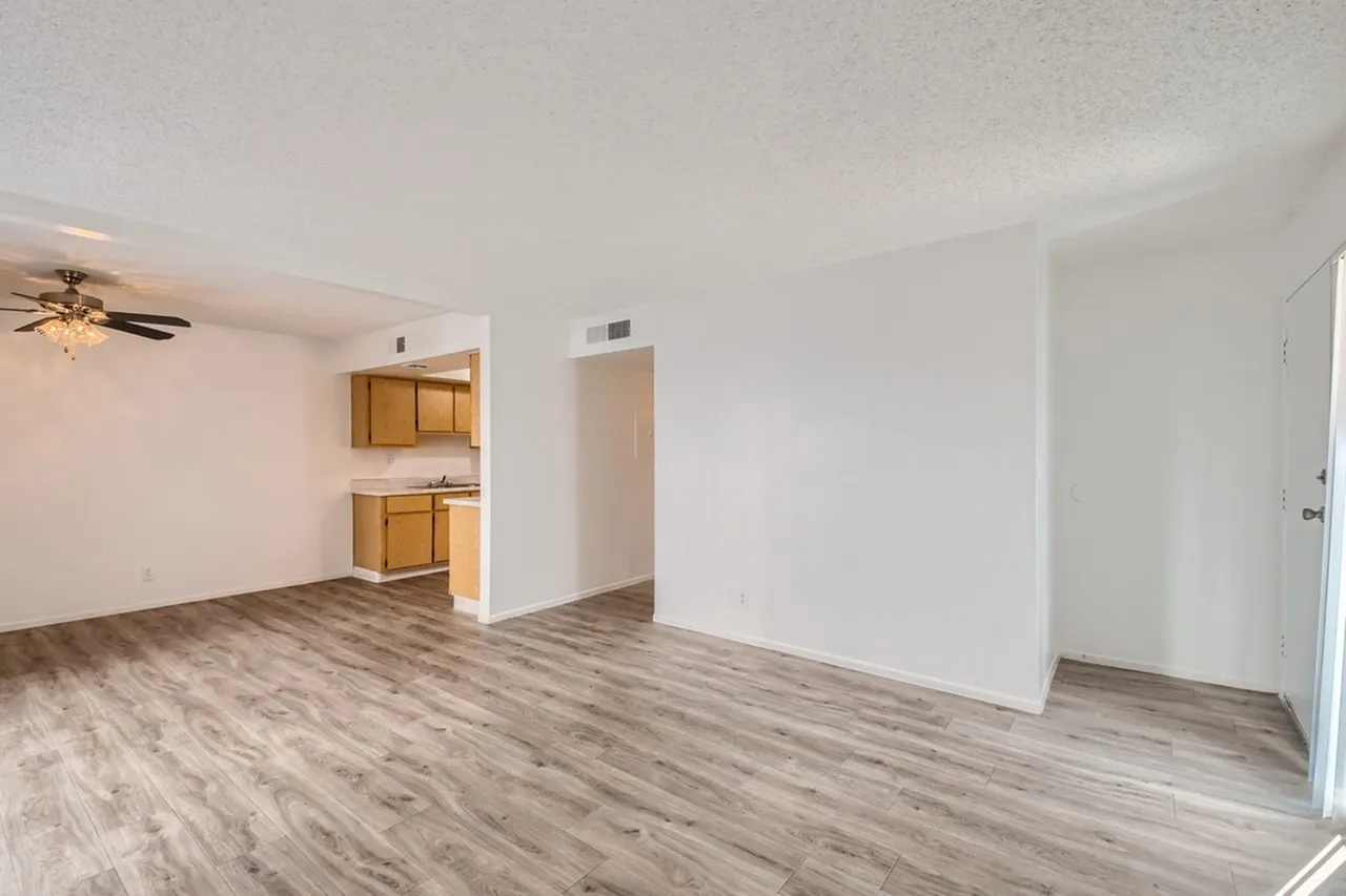 Living room with wood-style plank flooring, ceiling fan, kitchenette with oak cabinetry, entry door